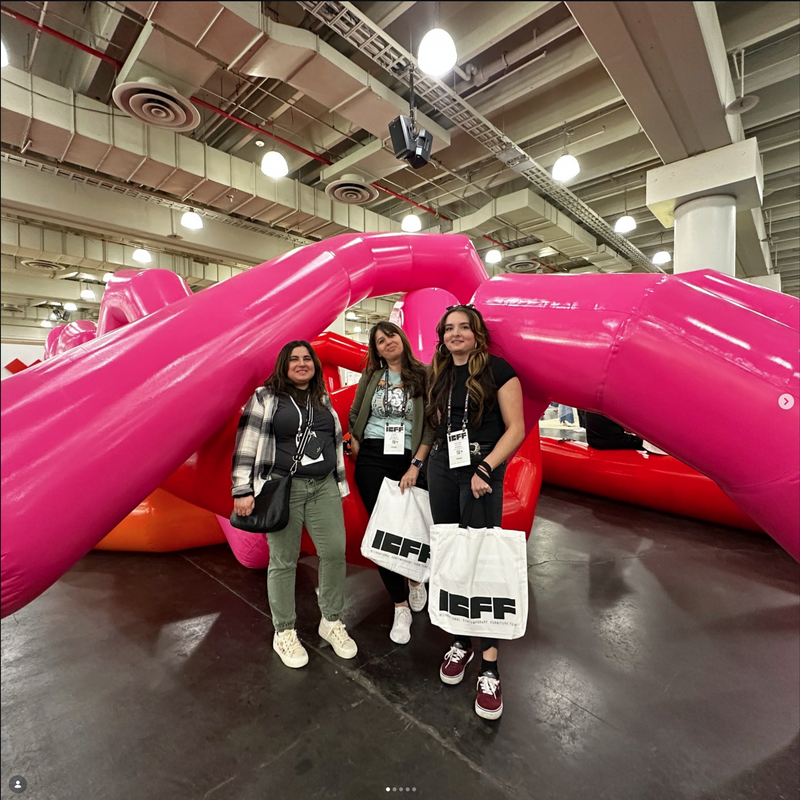 Three women standing in front of large inflatable slides with 'ICFF' bags in a warehouse setting.
